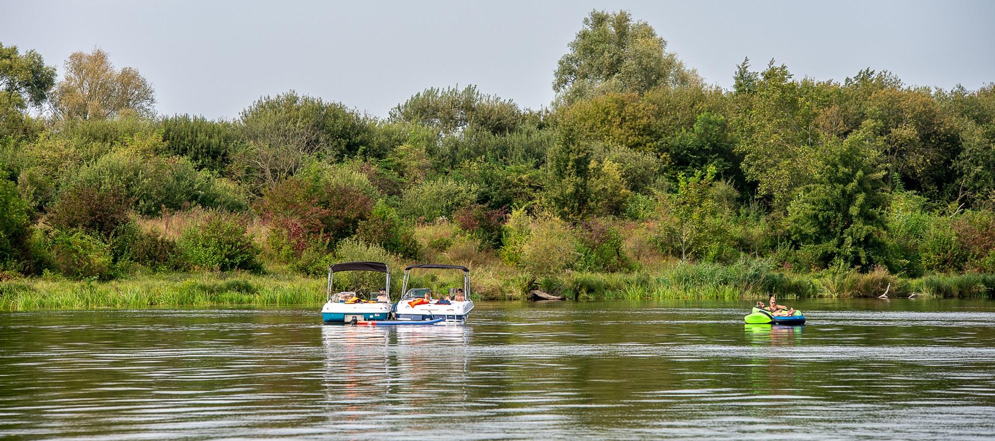 Twee motorboten en een opblaasbootje met mensen op een rustige rivier, omringd door groen en bomen.