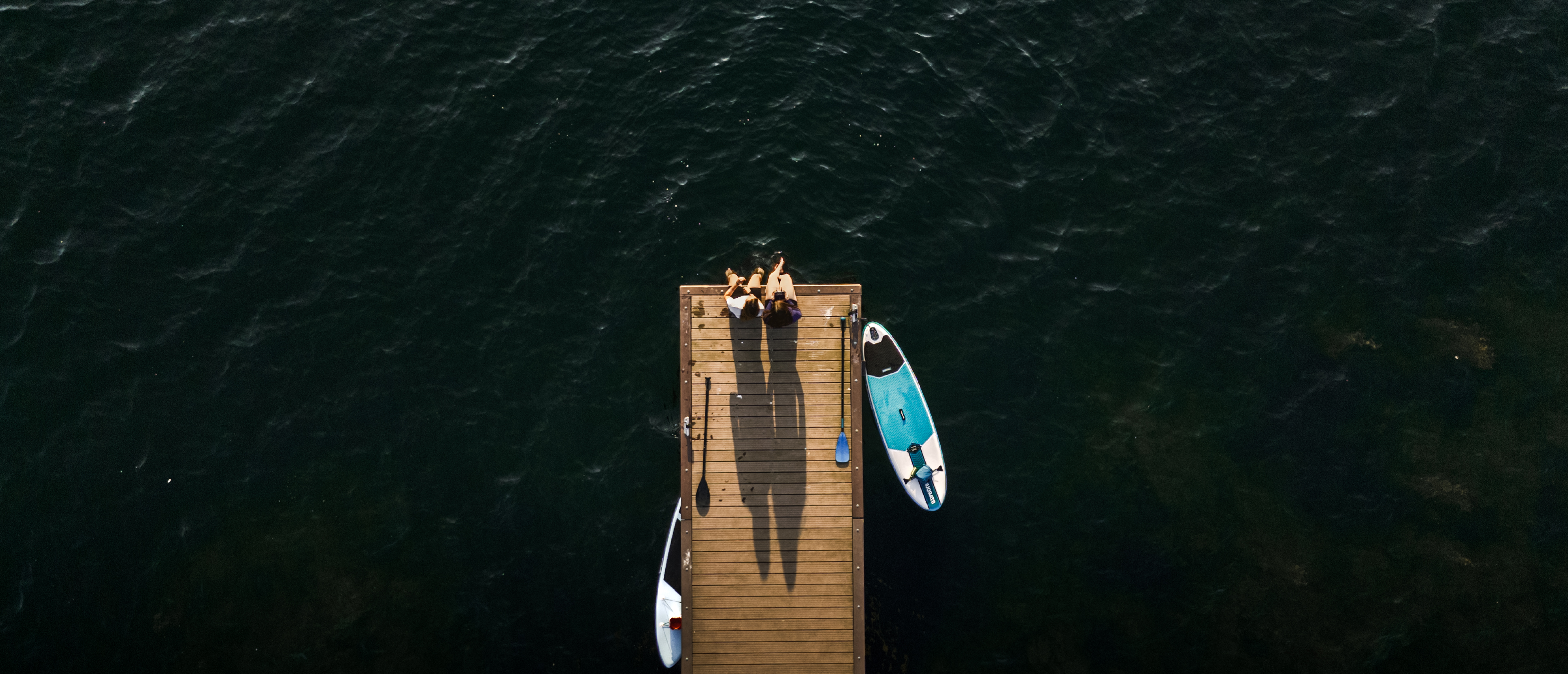Luchtfoto van twee mensen die aan het einde van een houten steiger zitten boven donker water, met aan weerszijden een SUP-board en peddels in het water en op de steiger.