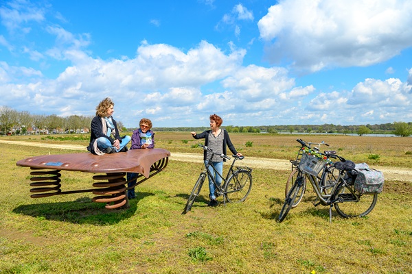 Twee vrouwen en een kind rusten uit bij een speels kunstwerk in het landschap, met uitzicht op velden.