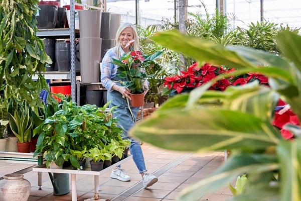 Vrouw in tuincentrum met bloempot in haar handen, omringd door groene planten en bloemen.