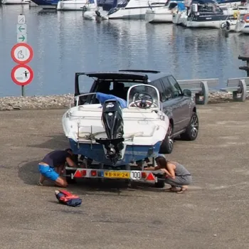 Twee personen maken een boottrailer gereed bij een trailerhelling, met een jachthaven op de achtergrond.